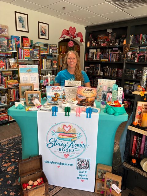 Stacey Loomis behind her table at No Shelf Control, with both Shelly's Adventures books and the activity book on stands, bookmarks, 3D-printed sea creatures, and a banner with the Stacey Loomis Books logo